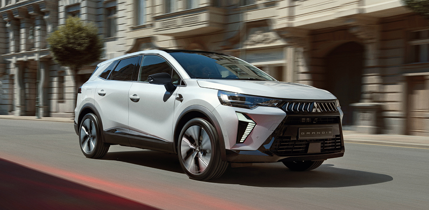 Geschäftskunden: Grandis Weißer Mitsubishi Grandis Kompakt-SUV fährt dynamisch eine sonnige Stadtstraße entlang, im Hintergrund Gebäude mit eleganter Fassade.