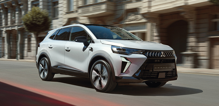 Geschäftskunden: Grandis Weißer Mitsubishi Grandis Kompakt-SUV fährt dynamisch eine sonnige Stadtstraße entlang, im Hintergrund Gebäude mit eleganter Fassade.