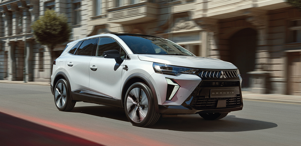 Geschäftskunden: Grandis Weißer Mitsubishi Grandis Kompakt-SUV fährt dynamisch eine sonnige Stadtstraße entlang, im Hintergrund Gebäude mit eleganter Fassade.
