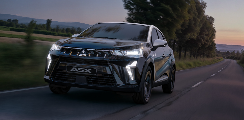 Mitsubishi ASX Black – dynamischer Auftritt in der Dämmerung Mitsubishi ASX Black Sondermodell fährt in der Dämmerung auf einer Landstraße, Front Seitenansicht mit eingeschalteten LED Scheinwerfern; die Szene betont den sportlich exklusiven Charakter der ASX Sondermodelle.