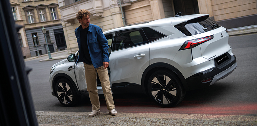 Mitsubishi Grandis Intro Edition in der Stadt Ein Mann in Jeansjacke steht neben einem weißen Mitsubishi Grandis Intro Edition vor einem Gebäude