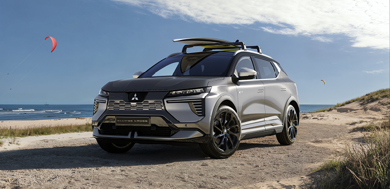 Eclipse Cross Zubehör Ein Mitsubishi Eclipse Cross steht auf einem sandigen Weg an der Küste, mit einem Surfbrett auf dem Dachträger; im Hintergrund Meer, Strand und bunte Kite-Segel am Himmel.